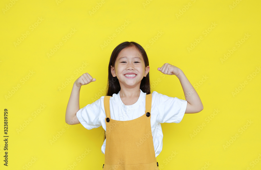 Strong asian little kid girl raising hands up and smiling. Portrait of happy child in dungarees isolated on yellow background. Strong asian little kid girl raising hands up and smiling. Portrait of happy child in dungarees isolated on yellow background.