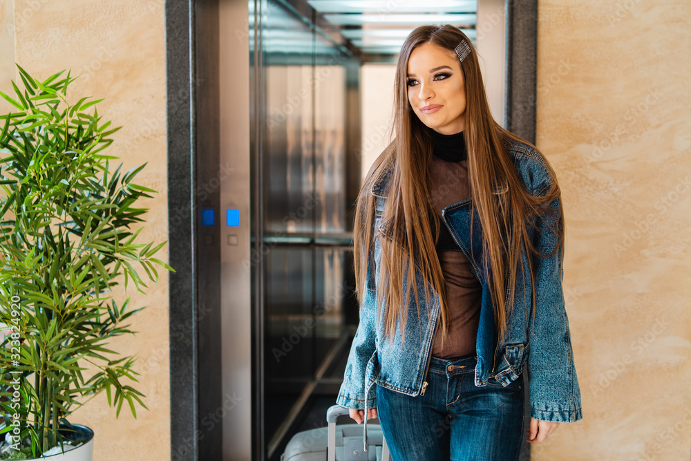 Portrait of young woman caucasian girl female tourist travel walking from the elevator of the hotel or motel looking to the camera while dragging the suitcase on vacation wearing jeans jacket