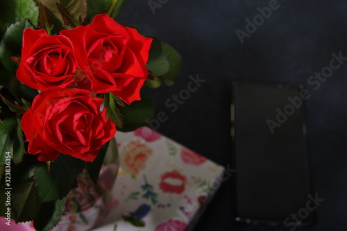 Bouquet red roses flower in glass vase on dark background and black smartphone no focus, gift box