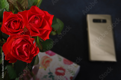 Bouquet red roses flower in glass vase on dark background and gold smartphone no focus, gift box