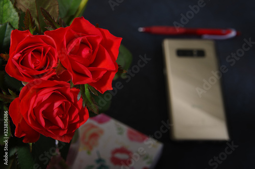 Bouquet red roses flower in glass vase on dark background and pen at the gold smartphone no focus, gift box