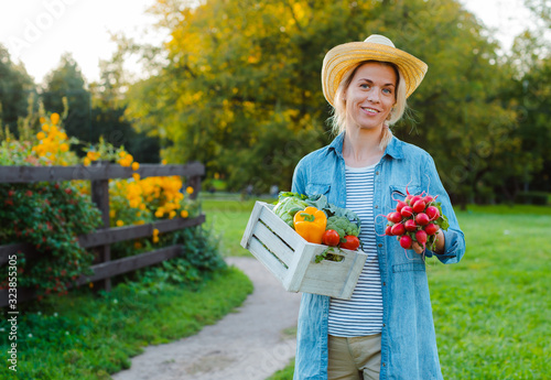 Young 30-35 years old beautiful Woman farmer in hat with box of fresh ecological vegetables on garden background at sunset.