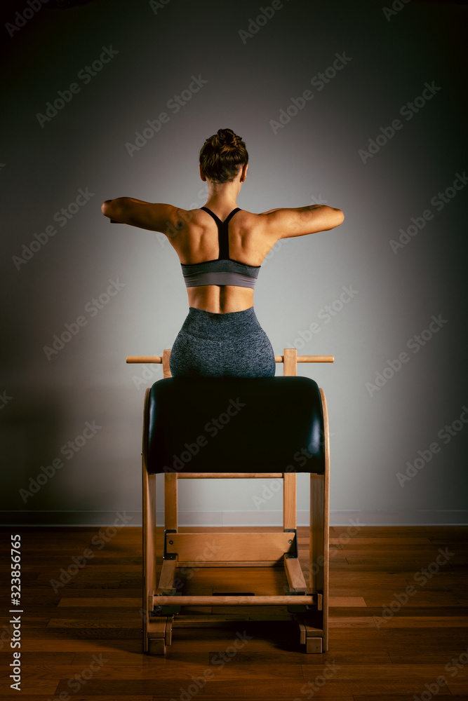 Fototapeta A young girl does Pilates exercises with a bed reformer, barrel machine tool. Beautiful slim fitness trainer on a background of a reformer gray, low key, light art. Fitness concept, healthy lifestyle.