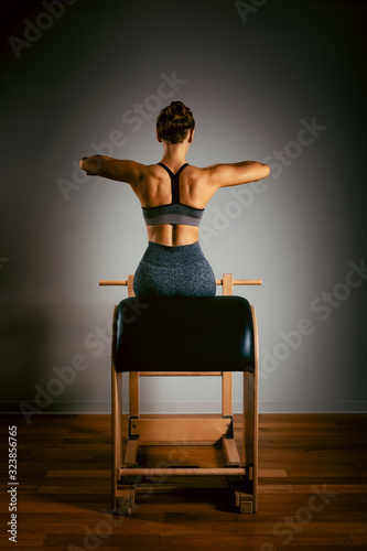 Fototapeta A young girl does Pilates exercises with a bed reformer, barrel machine tool. Beautiful slim fitness trainer on a background of a reformer gray, low key, light art. Fitness concept, healthy lifestyle.