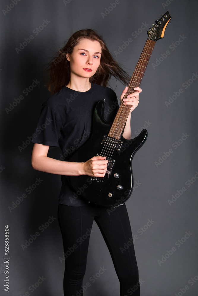 Portrait of a beautiful, young rock girl in black clothes with an electric guitar in her hands. Studio photo on a gray background. Model with clean skin.