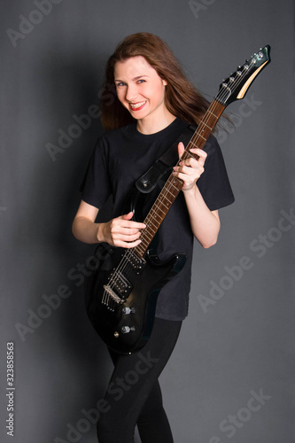 Portrait of a beautiful, young rock girl in black clothes with an electric guitar in her hands. Studio photo on a gray background. Model with clean skin.