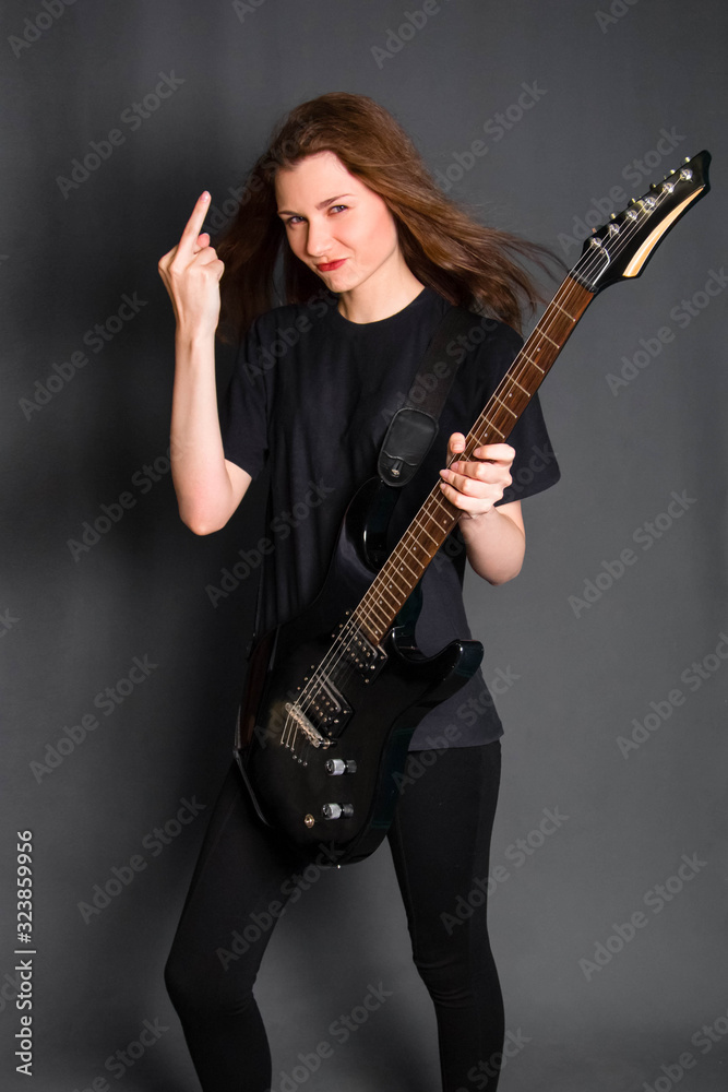 Angry rock girl in black clothes holds an electric guitar in her hands and shows the middle finger, gesture fuck off. Studio photo on a gray background. Angry rock girl in black clothes holds an electric guitar in her hands and shows the middle finger, gesture fuck off. Studio photo on a gray background.