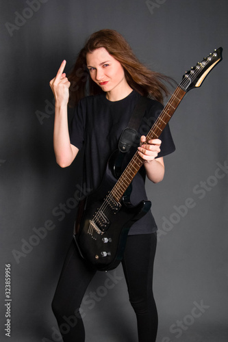 Angry rock girl in black clothes holds an electric guitar in her hands and shows the middle finger, gesture fuck off. Studio photo on a gray background.