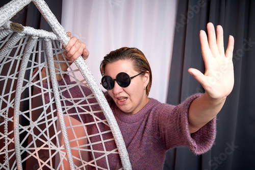 Ugly aged woman in a sweater and black glasses in a white wicker hanging chair in the room with curtains