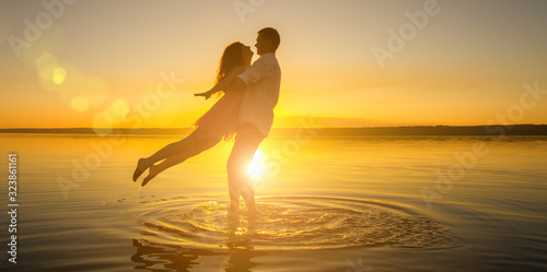 Young wedding couple is hugging in the water on summer beach. Beautiful sunset over the sea.Two silhouettes against the sun. Romantic love story. Man and woman in love in honeymoon trip.
