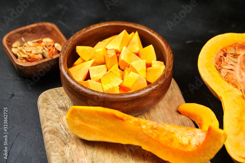 Butternut squash pieces, wooden bowl with pumpkin seeds, cutting board on black background, closeup. Cooking winter squash