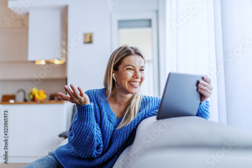 Картината върху платно Beautiful caucasian blond woman dressed in blue sweater sitting on sofa in living room and having video call over tablet