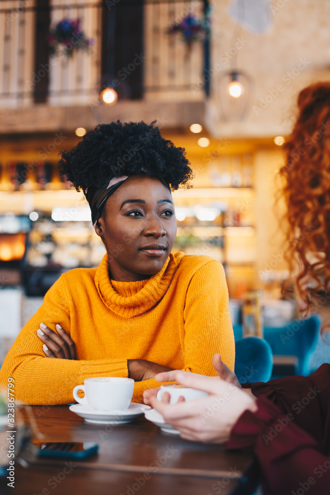 Two female friends sitting at the cafe drinking coffee, talking. The focus is on the African young lady. Two female friends sitting at the cafe drinking coffee, talking. The focus is on the African young lady.