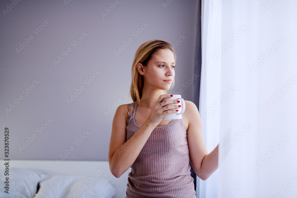 Attractive serious caucasian blond woman in pajamas standing next to window, looking trough it and holding mug with fresh coffee. Bedroom interior. Attractive serious caucasian blond woman in pajamas standing next to window, looking trough it and holding mug with fresh coffee. Bedroom interior.