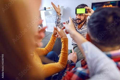 Group of cheerful playful friends playing charades with smart phone. Home interior.