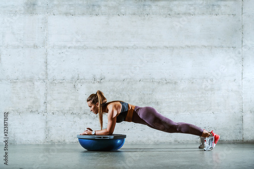 Side view of young muscular caucasian sporty brunette in sportswear with ponytail doing planks and leaning on bosu ball. Healthy lifestyle concept. photo