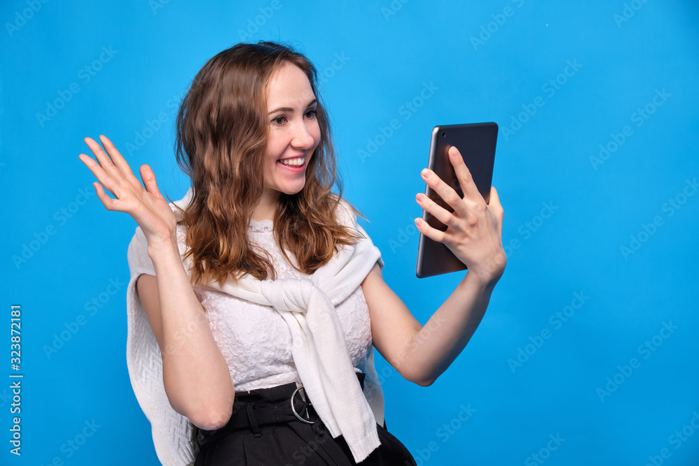 Fototapeta premium A brunette girl in casual clothes on a blue background sits and communicates via video link on a tablet.