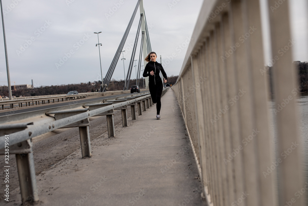 Young woman in black sports outfit running on the bridge in the city during day.