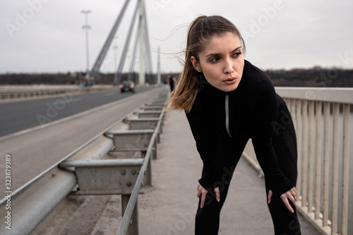 Young woman in black sports outfit resting after running on the bridge in the city.