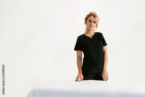 Portrait of young smiling friendly funny woman masseur in uniforme near massage table in Spa Salon. Physical Therapist In Medical Office. Beauty Treatment, Massage Therapy. White background isolated photo
