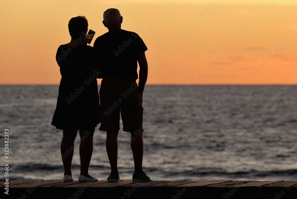 Silhouette of an older couple on the ocean coast as they take pictures of the sunset
