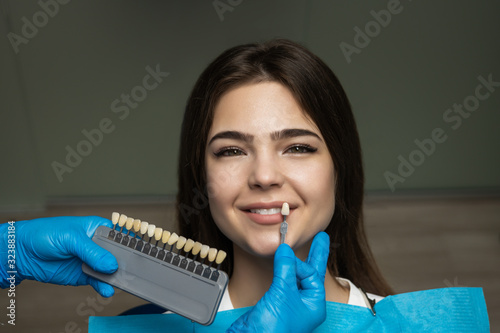 dentist in blue medical gloves applying sample from tooth enamel scale to happy woman patient teeth to choose right shade for teeth bleaching procedure