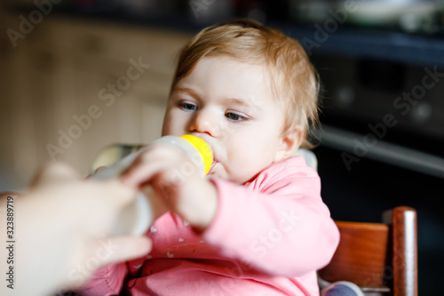 Cute adorable baby girl holding nursing bottle and drinking formula milk. First food for babies. New born child  sitting in chair of domestic kitchen. Healthy babies and bottle-feeding concept