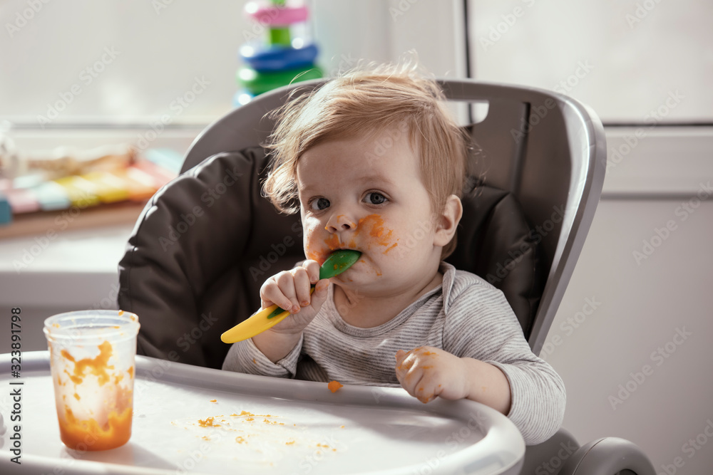 Beautiful baby in the child dining chair eat healthy food. Child eat vegetable puree. Lifestyle. Sunny day. Dirty face of happy kid. Portrait of a baby eating with a stained face. Beautiful baby in the child dining chair eat healthy food. Child eat vegetable puree. Lifestyle. Sunny day. Dirty face of happy kid. Portrait of a baby eating with a stained face.