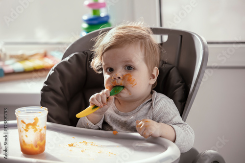 Beautiful baby in the child dining chair eat healthy food. Child eat vegetable puree. Lifestyle. Sunny day. Dirty face of happy kid. Portrait of a baby eating with a stained face.