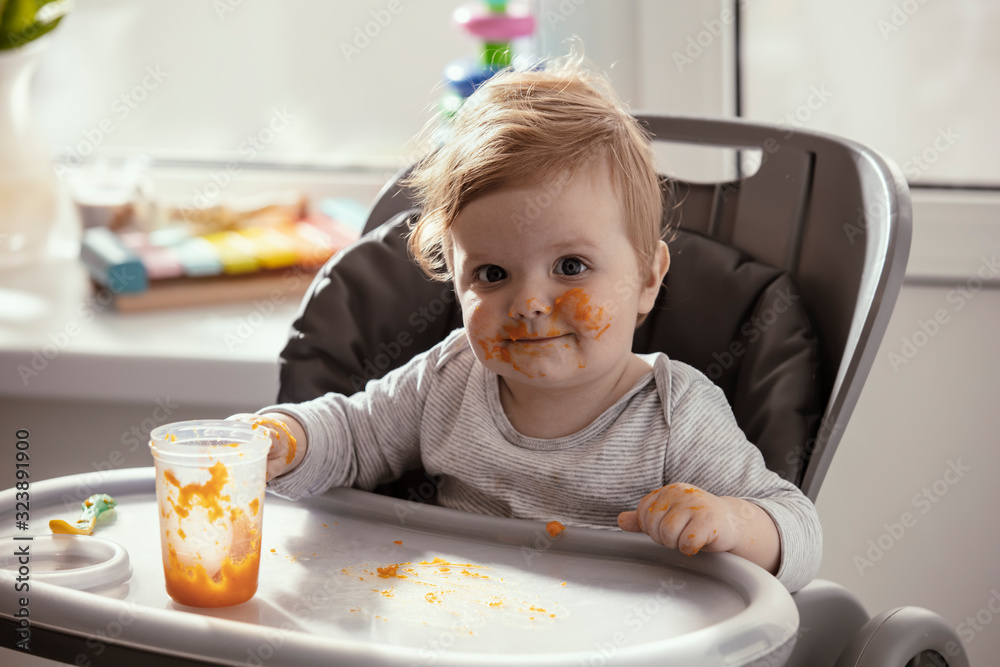 Beautiful baby in the child dining chair eat healthy food. Child eat vegetable puree. Lifestyle. Sunny day. Dirty face of happy kid. Portrait of a baby eating with a stained face. Beautiful baby in the child dining chair eat healthy food. Child eat vegetable puree. Lifestyle. Sunny day. Dirty face of happy kid. Portrait of a baby eating with a stained face.