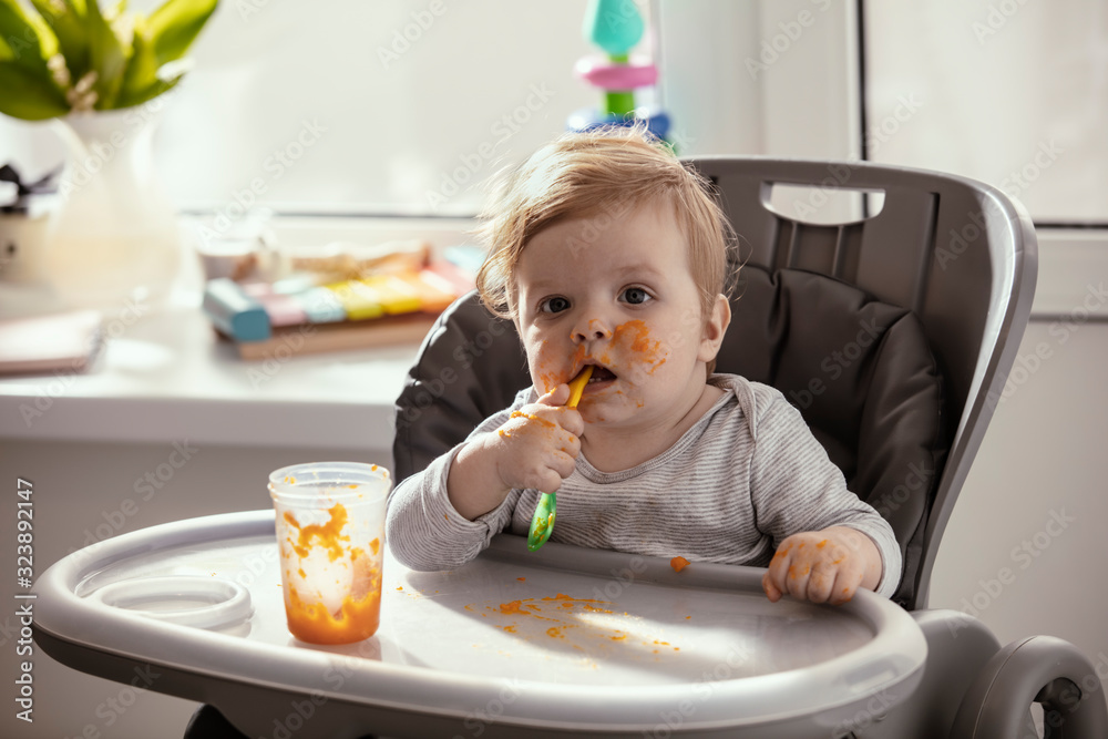 Beautiful baby in the child dining chair eat healthy food. Child eat vegetable puree. Lifestyle. Sunny day. Dirty face of happy kid. Portrait of a baby eating with a stained face. Beautiful baby in the child dining chair eat healthy food. Child eat vegetable puree. Lifestyle. Sunny day. Dirty face of happy kid. Portrait of a baby eating with a stained face.