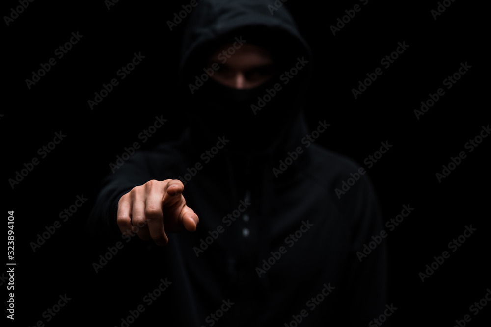 selective focus of hacker in hood and mask pointing with finger isolated on black