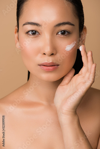 beautiful naked asian girl applying facial cream isolated on beige