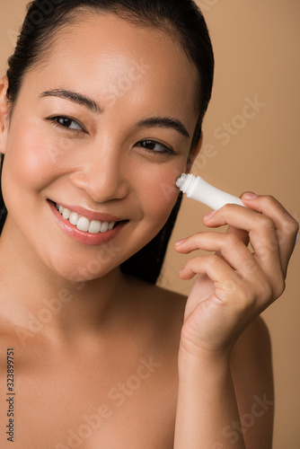 smiling beautiful naked asian girl using eye roller isolated on beige