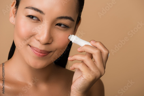 smiling beautiful naked asian girl using eye roller isolated on beige