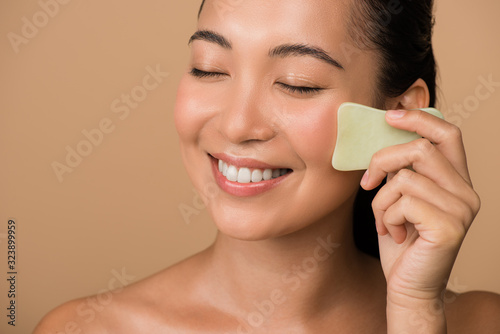 smiling beautiful naked asian girl with closed eyes using facial gua sha jade board isolated on beige photo