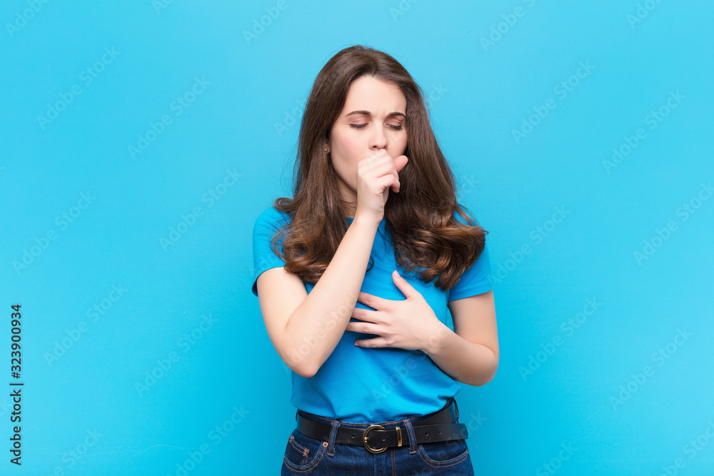 young pretty woman feeling ill with a sore throat and flu symptoms, coughing with mouth covered against blue wall - obrazy, fototapety, plakaty 