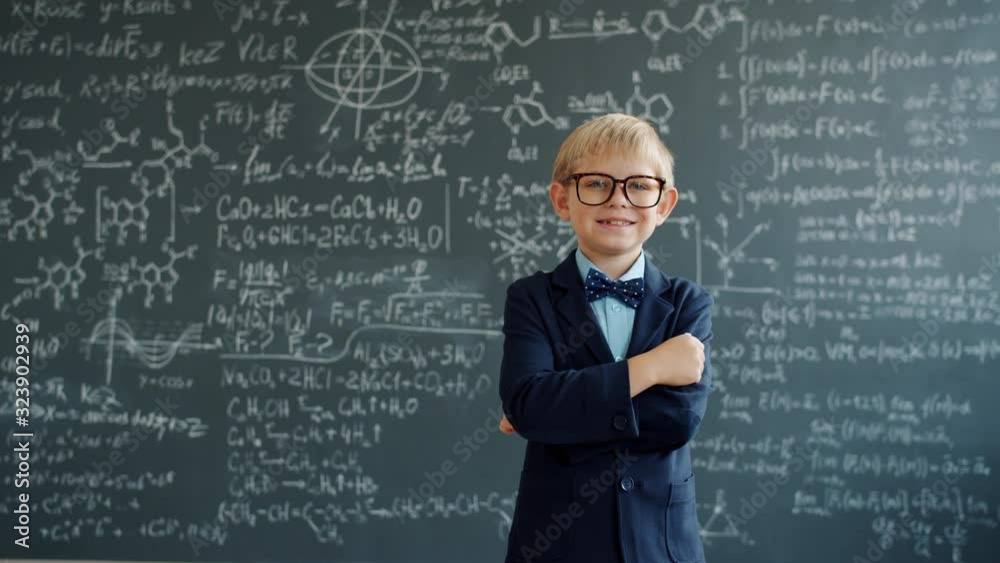 Happy kid in suit and glasses is standing in class with chalkboard in background with scientific formulas written on it. People, childhood and education concept. Happy kid in suit and glasses is standing in class with chalkboard in background with scientific formulas written on it. People, childhood and education concept.