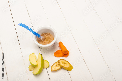 High angle view of baby food in bowl with spoon and sliced fruits and carrot on white wooden surface