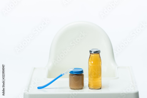 Jar of baby food with spoon and bottle of apple juice on table of feeding chair isolated on white