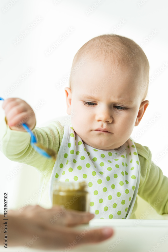 Selective focus of baby holding spoon near jar with vegetable puree and mother on white background Selective focus of baby holding spoon near jar with vegetable puree and mother on white background