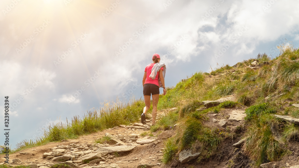 Girl young tourist in a pink t-shirt is walking along a mountain trail uphill against a background of clouds.