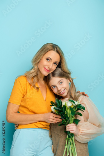 cheerful mother smiling near daughter with tulips isolated on blue