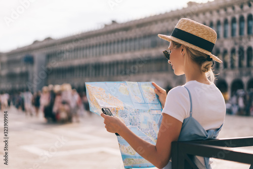 Caucasian female tourist in trendy apparel standing at popular Venezian San Marco Square, millennial woman with mobile technology and paper map visiting romantic city during solo journey to Italy