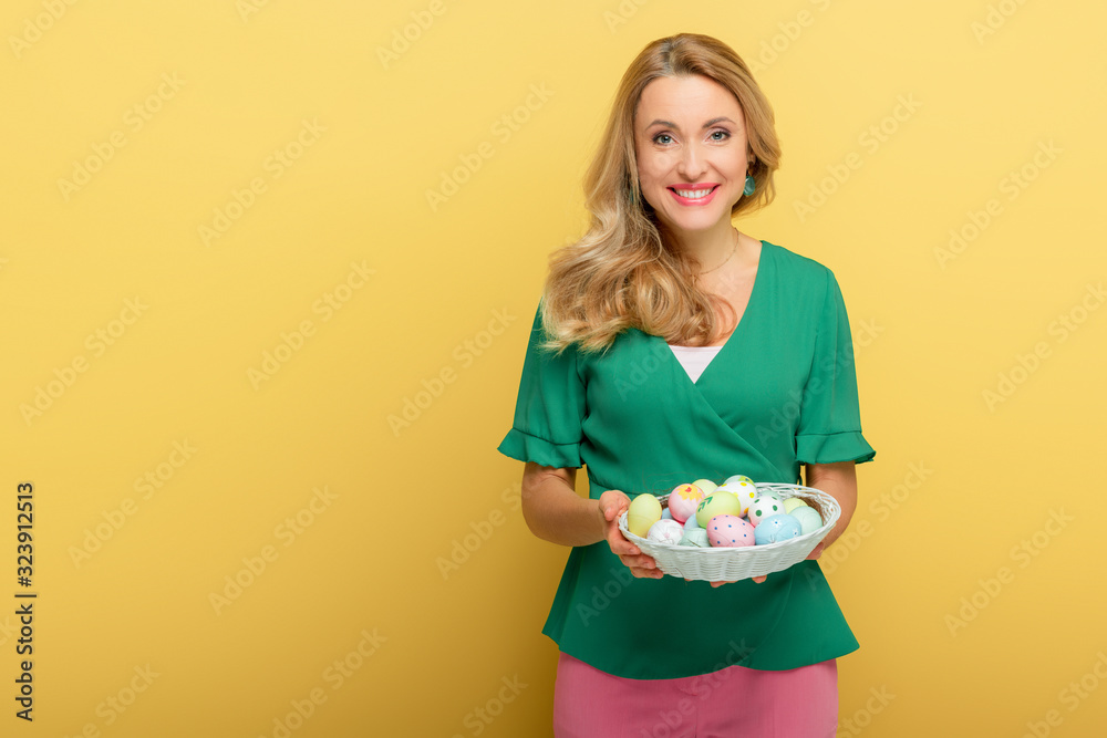 cheerful woman holding easter eggs in basket isolated on yellow