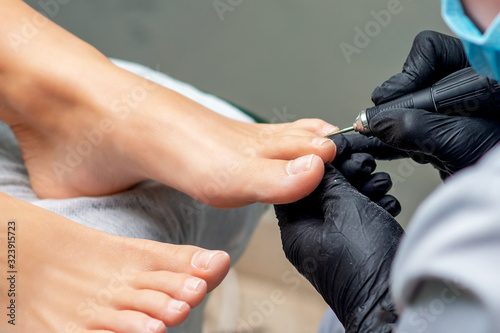 Pedicurist's hands in gloves with special tool doing pedicure of female nails on feet. Foot treatment in spa salon, close up.