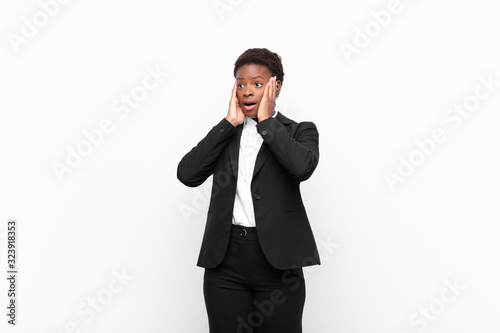 young pretty black womanfeeling happy, excited and surprised, looking to the side with both hands on face against white wall photo