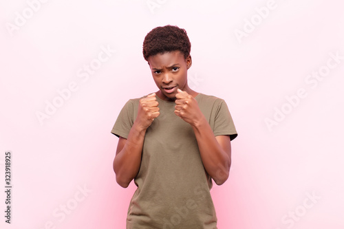 young pretty black womanlooking confident, angry, strong and aggressive, with fists ready to fight in boxing position against pink wall photo