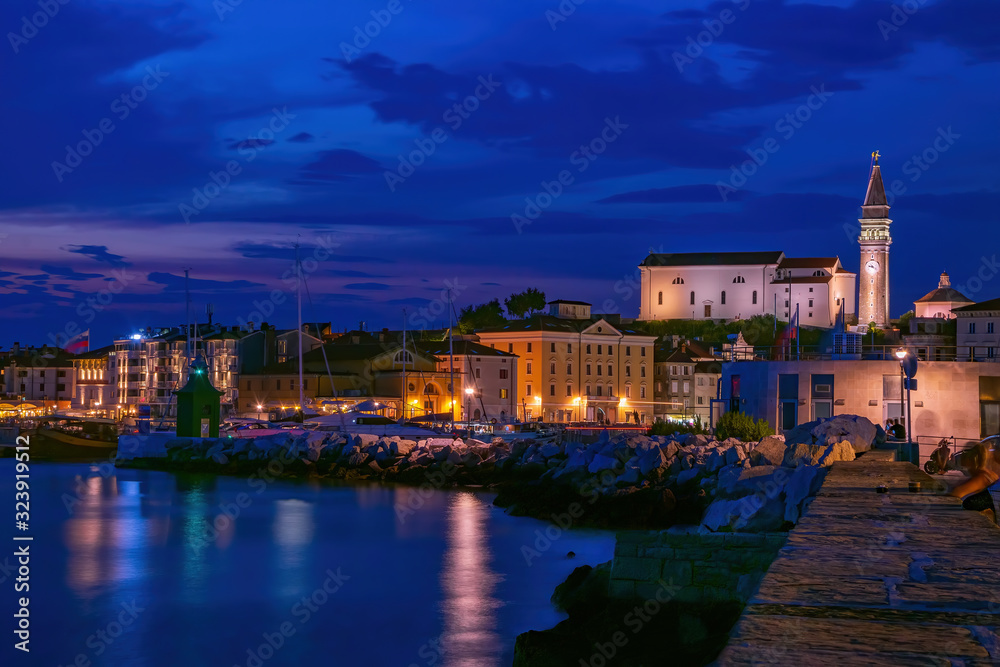 Picturesque nightscape of coastline of Adriatic sea with illuminated houses and lighthouse at twilight, Piran, Slovenia