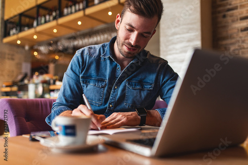 Freelancer working at cafe sitting at the table using laptop and writes in a notebook.